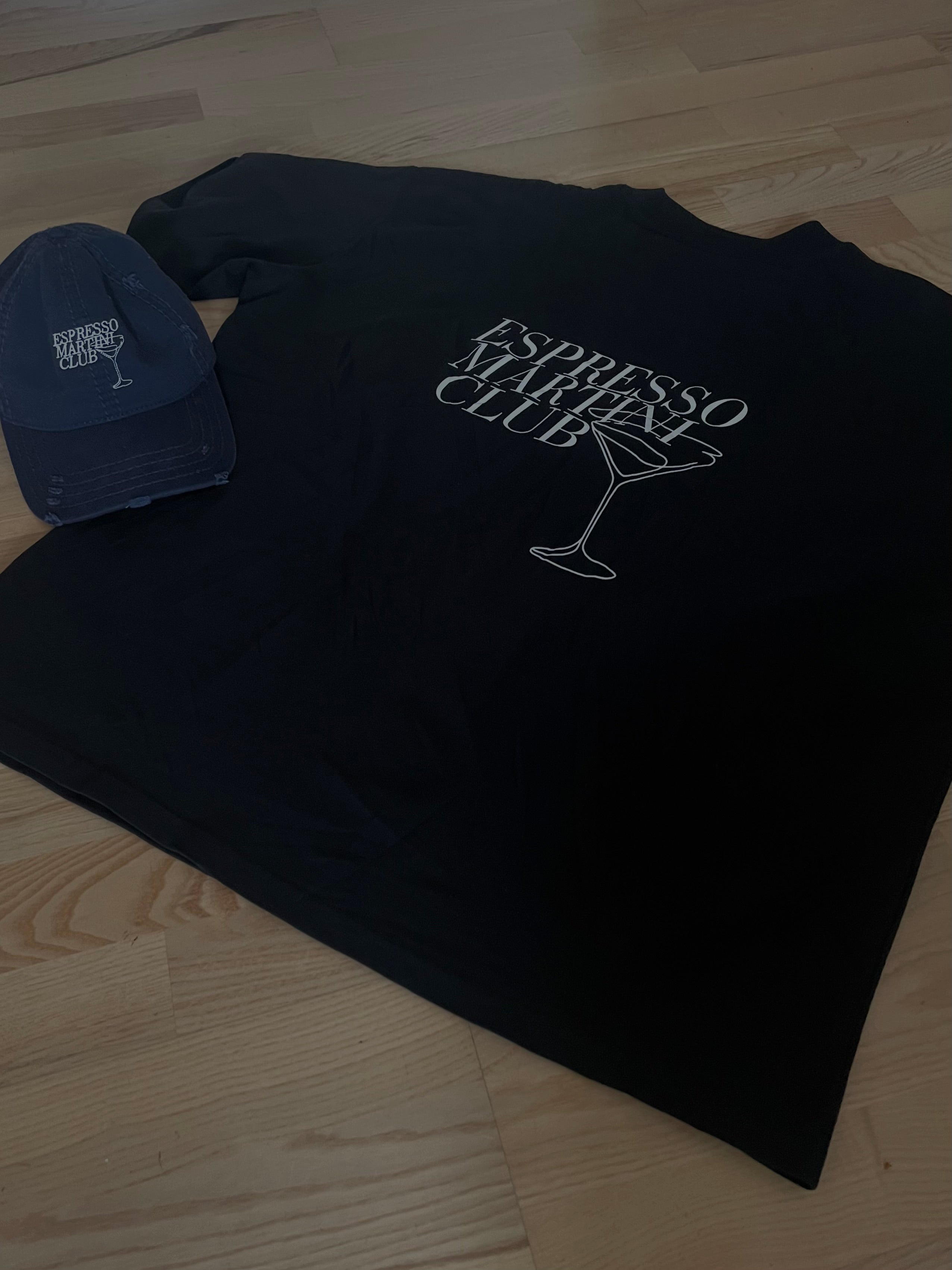 Black sweatshirt with 'Espresso Martini Club' text and logo on a wooden floor, next to a blue cap.